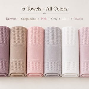 May include: Six rolled towels featuring a waffle-textured pattern and a smooth band. The text reads 6 Towels &ndash; All Colors, Damson + Cappuccino + Pink + Gray + White + Powder. The colors are dark purple, tan, light pink, light gray, white, and pale pink.