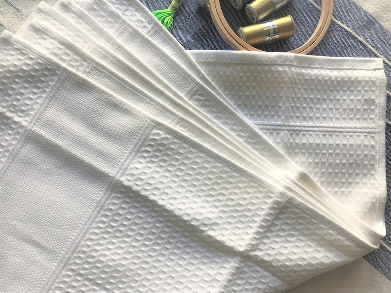 May include: A stack of white cotton dish towels with a waffle weave pattern. The towels are folded in a fan shape.