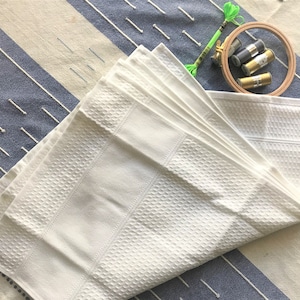 May include: A white waffle weave towel folded in a fan shape on a blue and white striped surface. There are three spools of embroidery floss and a wooden embroidery hoop in the background.