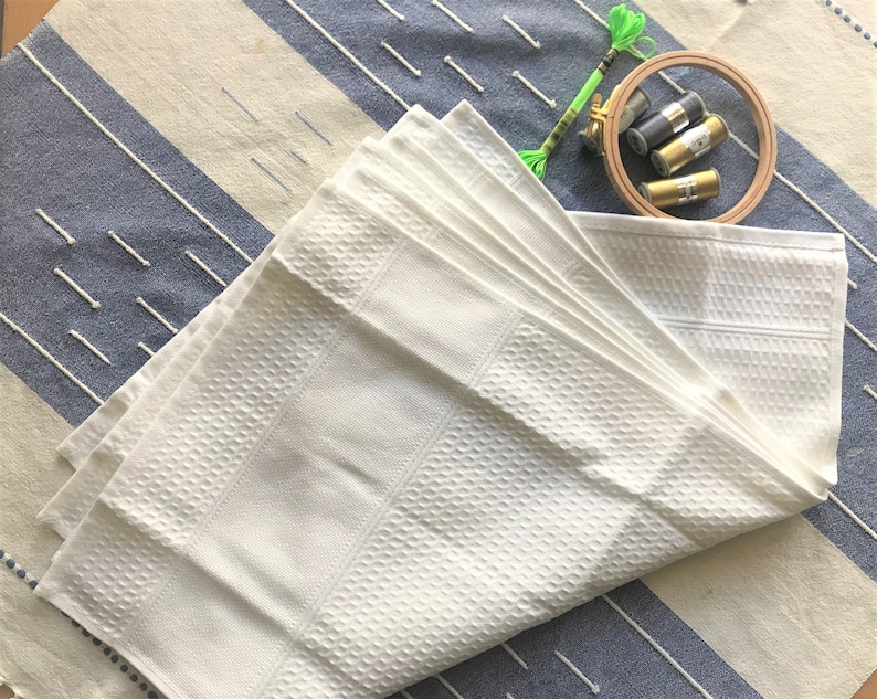 May include: A white waffle weave towel folded in a fan shape on a blue and white striped surface. There are three spools of embroidery floss and a wooden embroidery hoop in the background.