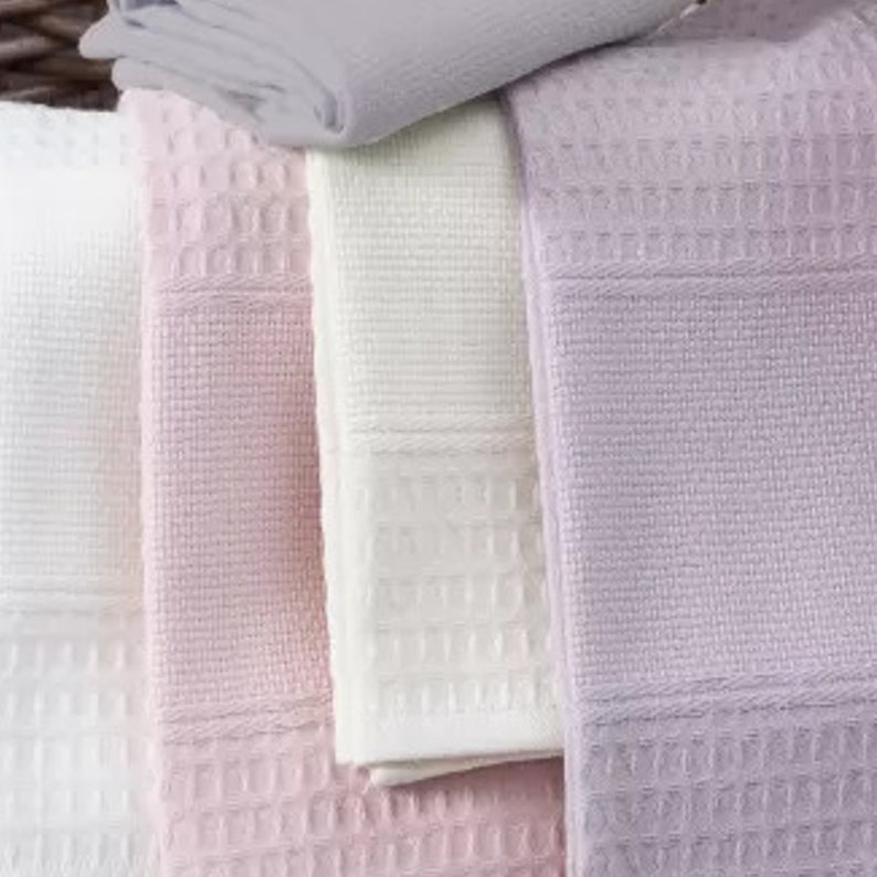 May include: A collection of folded waffle-weave towels in shades of white, light pink, lavender, and light gray. Each towel features a distinct grid-like texture and a horizontal band of tighter weave detail along the bottom edge of the fabric.