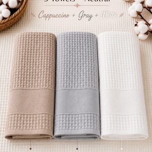 May include: A set of three waffle-weave towels in cappuccino, gray, and white. The text reads 3 Towels - Neutral, Cappuccino + Gray + White, with individual labels for Cappuccino, Gray, and White. Each towel features a textured grid pattern.