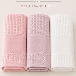 May include: Three rolled towels featuring a waffle-weave texture. The set includes one pink, one powder, and one white towel. Text at the top reads: 3 Towels &ndash; Soft Colors, Pink + Powder + White. The towels have a consistent grid pattern.