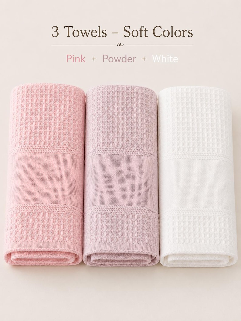 May include: Three rolled towels featuring a waffle-weave texture. The set includes one pink, one powder, and one white towel. Text at the top reads: 3 Towels &ndash; Soft Colors, Pink + Powder + White. The towels have a consistent grid pattern.