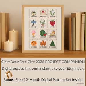 May include: A framed cross-stitch sampler featuring monthly icons like a coffee cup, heart envelope, and strawberry. The sampler includes the months January through December. The frame is light brown and sits on a wooden surface.