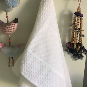 May include: A white waffle weave hand towel hanging on a hook. The towel has a textured pattern and is folded in half.