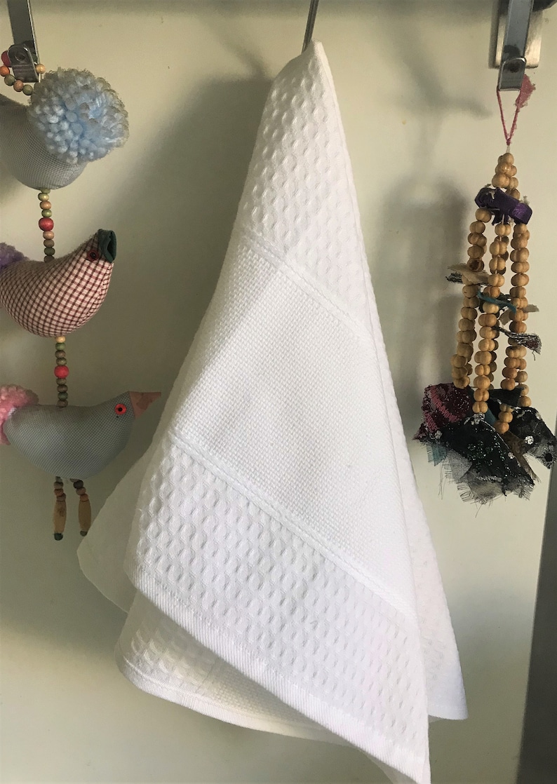 May include: A white waffle weave hand towel hanging on a hook. The towel has a textured pattern and is folded in half.