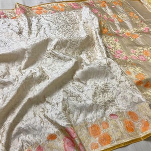 Puede incluir: Un textil color crema con una intrincada superposición de encaje blanco. La tela presenta un estampado floral en tonos de rosa y naranja a lo largo del borde. El textil tiene un ribete dorado.