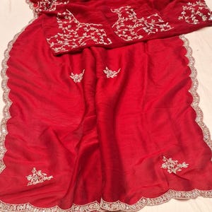 May include: A red silk saree with a scalloped border and silver embroidery. The embroidery features floral designs and is scattered throughout the saree.