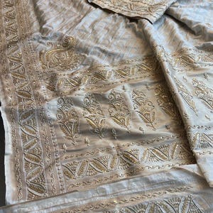 Könnte beinhalten: Ein weißer Seiden-Sari mit goldener Stickerei und Pailletten. Der Sari hat ein traditionelles indisches Design mit floralen Motiven und Paisley-Mustern.