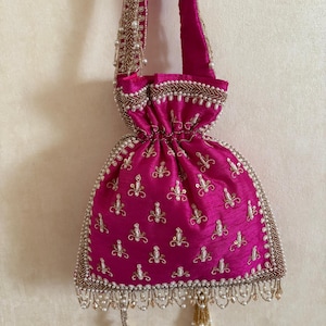 May include: A magenta drawstring bag with detailed gold and pearl adornments. The bag has a beaded handle and a decorative border with hanging pearl tassels. The fabric has a slight sheen, and the design includes embroidered floral patterns.