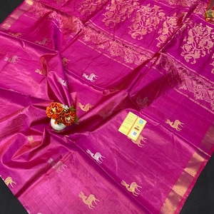 May include: A vibrant pink silk saree with gold floral and animal embroidery. The saree features intricate patterns and a shimmering texture. The border is trimmed with a thin gold band. A small vase of flowers sits on the saree.