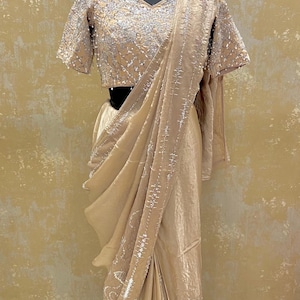 Peut inclure: Un sari beige avec des broderies et des ornements argentés complexes. Le sari est drapé sur un mannequin, mettant en valeur le travail détaillé du tissu. Le chemisier présente des manches courtes et une encolure carrée, également ornée d'accents argentés. L'ensemble est élégant et formel.