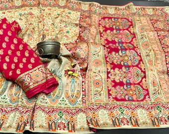 May include: A vibrant textile display featuring a fuchsia blouse with a paisley pattern and a decorative border. The fabric is draped over a cream-colored textile with intricate floral and paisley designs. A small metal container rests on the fabric.