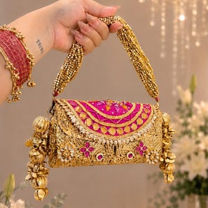 May include: A gold-toned metal clutch bag featuring an intricate filigree pattern, accented with vibrant fuchsia fabric panels, embedded mirrors, and small pink stones. The bag includes a beaded handle and decorative gold-toned metal floral charms.