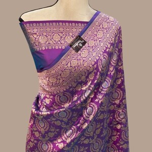May include: A purple and blue silk saree with a gold brocade design. The saree has a border with a floral pattern. The saree is draped on a mannequin.
