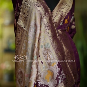 Puede incluir: Un sari de seda Banarasi Mashru de color morado con intrincados bordados de oro y plata. El sari presenta un diseño tradicional con motivos florales y un borde con un patrón de paisley. El sari está hecho con un tejido fino y tiene una sensación suave y lujosa. El sari es perfecto para ocasiones especiales y festivales.