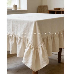 Puede incluir: Mantel de lino color crema con borde con volantes. El mantel está sobre una mesa de madera, con el texto "Brokenflower Gifts" visible en la parte delantera.