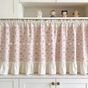 Peut inclure: Un rideau de cuisine rose à motifs floraux avec des volants blancs en haut et en bas. Le rideau présente un motif répété de petites fleurs et le texte "Broken Flower Gifts". Le rideau est suspendu devant un meuble blanc.