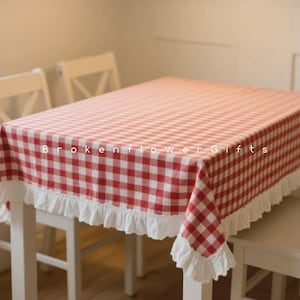 Puede incluir: Un mantel a cuadros rojos y blancos con un borde con volantes blancos cubre una mesa rectangular. El mantel está drapeado sobre una mesa de madera blanca, con dos sillas blancas visibles en el fondo. El texto "Brokenflower Gifts" está impreso en el mantel.