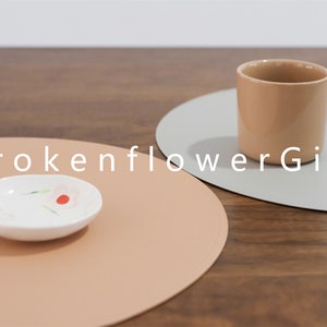 May include: Two round placemats, one light brown and one light gray, with a small white dish with red and pink flowers on the brown placemat and a light brown ceramic mug on the gray placemat. The text "BrokenflowerGift" is printed in white on the table surface.
