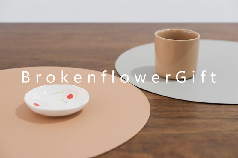 May include: Two round placemats, one light brown and one light gray, with a small white dish with red and pink flowers on the brown placemat and a light brown ceramic mug on the gray placemat. The text "BrokenflowerGift" is printed in white on the table surface.