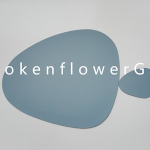 May include: Two blue leather placemats in the shape of an abstract leaf. The larger placemat is in the shape of a leaf with a rounded tip. The smaller placemat is a rounded oval shape. The text "BrokenflowerGift" is printed in white on a white background.