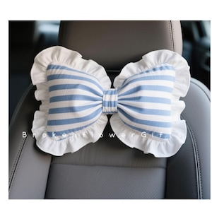 May include: A bow-shaped car headrest pillow with blue and white stripes and white ruffled edges. The pillow is placed on a gray car seat. The text "Broken Flower Gifts" is visible.
