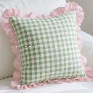 Pode incluir: Uma almofada quadrada com um padrão xadrez verde e branco, emoldurada por uma borda rosa com babados. A almofada está sobre uma superfície branca. O texto "Brokenflower Gifts" é visível na almofada.