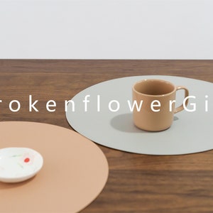 May include: Two round placemats, one light brown and one light gray, with a light brown ceramic mug on the gray placemat. The text "BrokenflowerGift" is printed in white on the table surface.