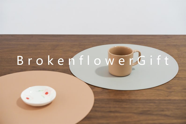 May include: Two round placemats, one light brown and one light gray, with a light brown ceramic mug on the gray placemat. The text "BrokenflowerGift" is printed in white on the table surface.