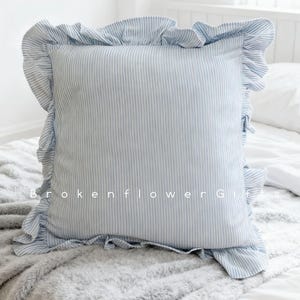 Puede incluir: Un cojín cuadrado con un diseño de rayas azules y blancas y un borde con volantes. El cojín está sobre una cama con una manta gris. El texto "Brokenflower Gifts" es visible.