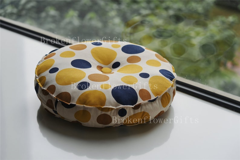 Op de afbeelding: Rond kussen met een witte stoffen hoes en een patroon van gele, bruine en blauwe stippen. Het kussen heeft een knoop in het midden.