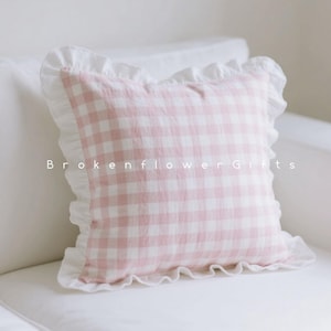 May include: A square cushion with a pink and white gingham design. It features a white, ruffled border. The text "Brokenflower Gifts" is visible. The cushion is placed on a white surface.