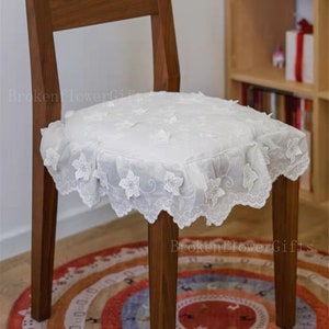 May include: A white lace chair cushion with a scalloped edge and floral embroidery. The cushion is placed on a wooden chair with a brown finish. The chair is sitting on a colourful rug with a geometric pattern.