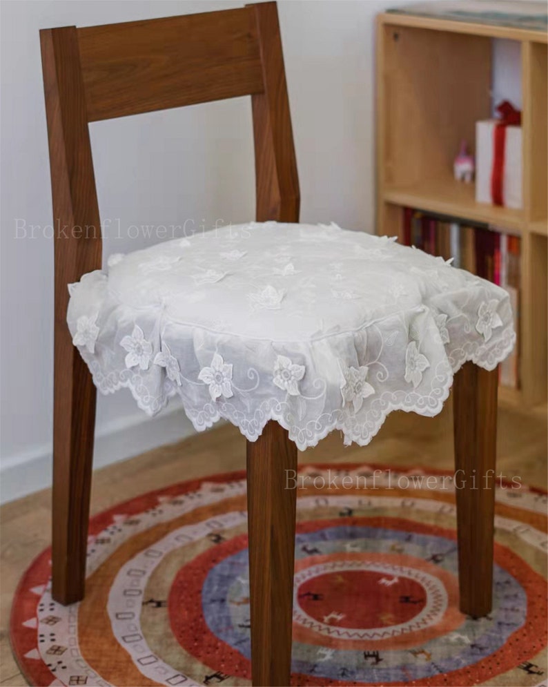 May include: A white lace chair cushion with a scalloped edge and floral embroidery. The cushion is placed on a wooden chair with a brown finish. The chair is sitting on a colourful rug with a geometric pattern.