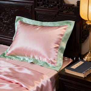 May include: A pink satin pillow with a light green border rests on a bed with matching pink sheets. The pillow is centered on the bed, with the text "Broken Flower Gifts" visible at the bottom.