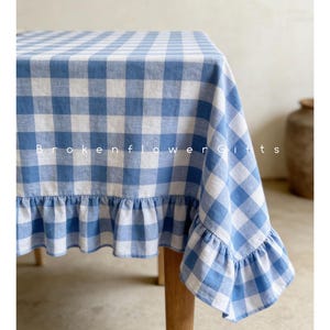 Op de afbeelding: Een blauw-wit geruite tafelkleed met een gefranjerde rand. Het tafelkleed is over een houten tafel gedrapeerd, met de tekst "Brokenflower Gifts" zichtbaar aan de voorkant. De stof heeft een klassieke, landelijke uitstraling.