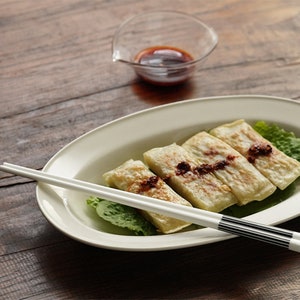 May include: Four golden brown dumplings on a white oval plate with a bed of green lettuce. A pair of white chopsticks with black stripes are resting on the plate. A small glass bowl with red sauce is in the background.