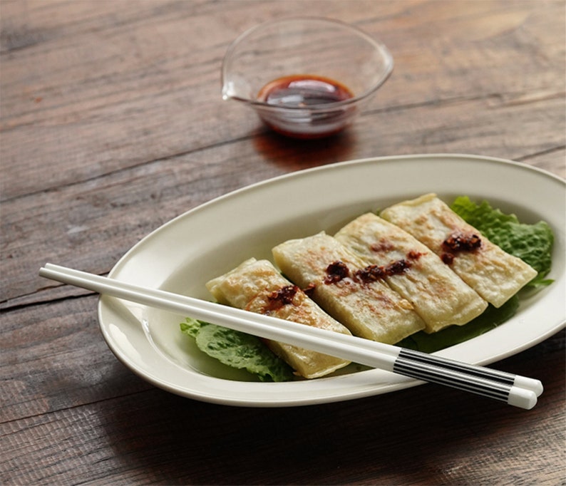 May include: Four golden brown dumplings on a white oval plate with a bed of green lettuce. A pair of white chopsticks with black stripes are resting on the plate. A small glass bowl with red sauce is in the background.