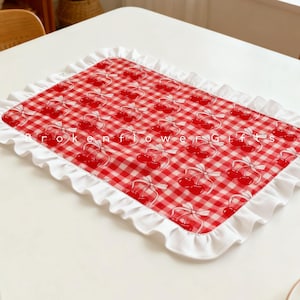 May include: A rectangular red and white checkered placemat with a repeating cherry pattern and a white ruffled edge. The text "Broken Flower Gifts" is printed on the placemat.
