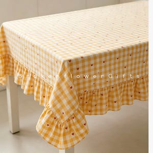 May include: A rectangular tablecloth featuring a yellow and white gingham check pattern with small red heart icons scattered across the fabric. The tablecloth has a ruffled edge and the text BrokenflowerGifts is printed across the center.