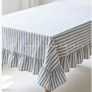 May include: A rectangular tablecloth with blue and white vertical stripes and a ruffled edge. The tablecloth is made of a light-colored fabric and is draped over a wooden table. The text "Broken Flower Gifts" is visible on the tablecloth.