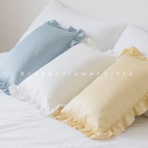 May include: Three rectangular pillows with frilled edges in pale blue, white, and yellow, arranged on a white bed. The pillows have a soft, textured appearance. The text "Brokenflower Gifts" is printed on the pillows.