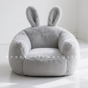 Peut inclure: Un fauteuil gris en peluche pour enfants en forme de lapin. Le fauteuil a une assise arrondie, des accoudoirs et deux oreilles de lapin dressées. Le texte "Brokenflower Gifts" est imprimé sur le devant du fauteuil.