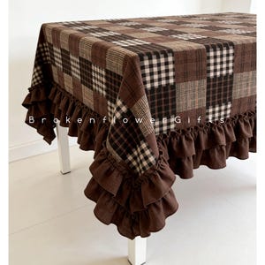 May include: A brown and cream plaid tablecloth with a ruffled edge. The tablecloth is covering a white table. The text "Brokenflower Gifts" is printed on the tablecloth.