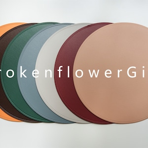 May include: Set of six round leather placemats in various colours, including orange, brown, green, blue, grey, and pink. The placemats are arranged in a row and have a stitched edge.