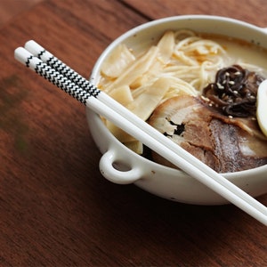 May include: A white bowl with a handle filled with ramen noodles, pork belly, a soft boiled egg, and bamboo shoots. A pair of white chopsticks with black and white patterned accents are placed on top of the bowl.