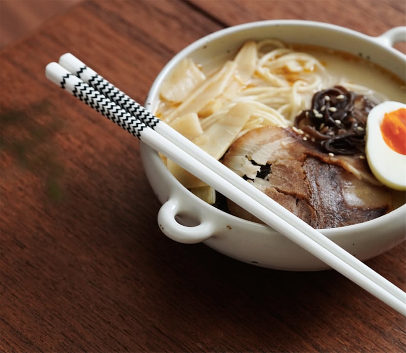 May include: A white bowl with a handle filled with ramen noodles, pork belly, a soft boiled egg, and bamboo shoots. A pair of white chopsticks with black and white patterned accents are placed on top of the bowl.