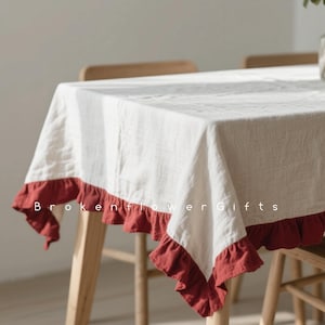 Op de afbeelding: Een witte rechthoekige tafelkleed met een gefranjerde, bordeauxrode rand. Het tafelkleed is gedrapeerd over een houten tafel, met de tekst "Brokenflower Gifts" zichtbaar.